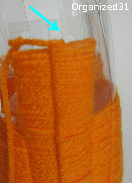 a blue arrow pointing to holes punched in the top of a plastic bottle with orange yarn wrapped around it