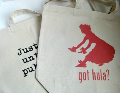 2 tote bags, the top one has an image of a lady hula dancing with text reading got hula?, the bottom one if covered by the other tote so the only text you can see says Just unt pub
