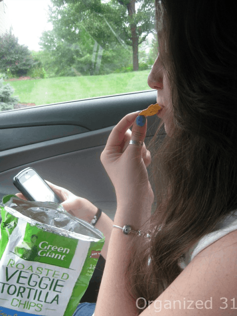 a teen eating a chip from a bag of chips while sitting in a car holding a cell phone