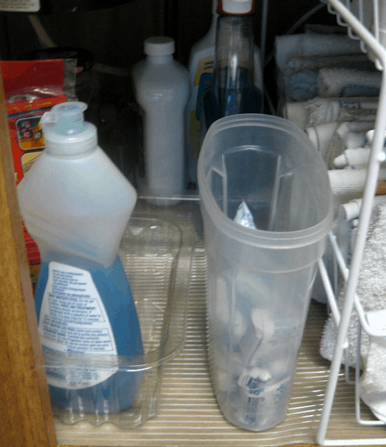 dishwasher tabs in frosted container with no lid next to cleaning supplies under kitchen sink.
