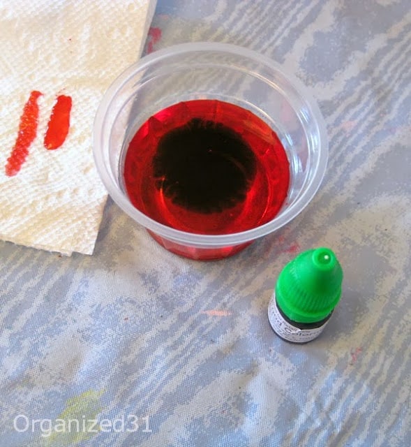 clear cup of red liquid with green food coloring bottle and red stained paper towel