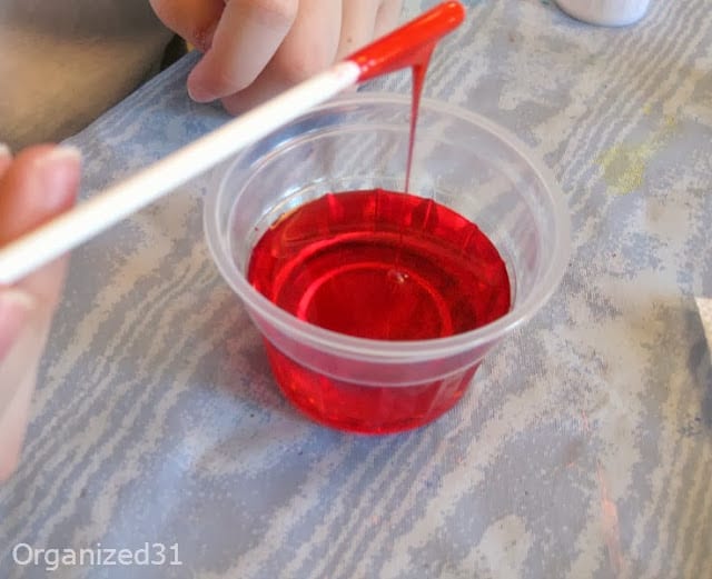 hand holding white stirring stick with thick red liquid dropping into cup of red liquid