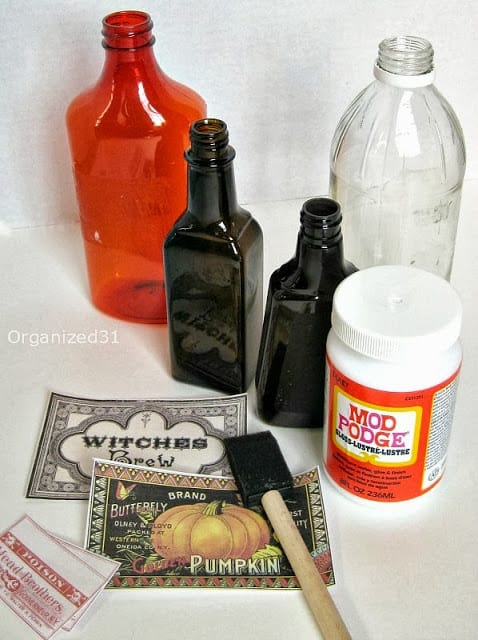 orange bottle, clear bottle and 2 dark brown bottles, bottle of decoupage glue, paper labels and sponge brush on white table
