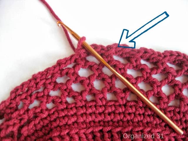 close-up of gold crochet needle and border of maroon crocheted cozy to show particular stitch.