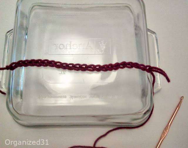 glass casserole dish with one maroon crocheted chain laying across top and gold crochet needle.
