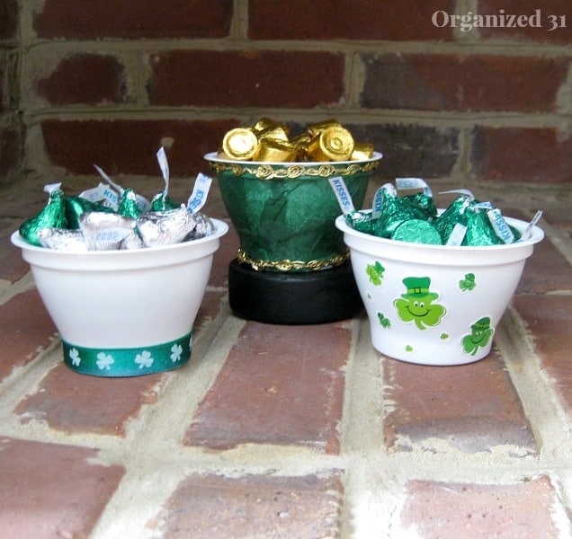 2 small white St. Patrick's decorated cups filled with green and silver candy with green cup in the background filled with gold foil wrapped candy on a brick background