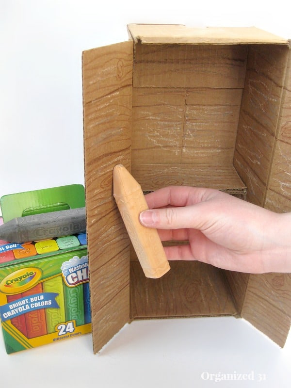 a hand holding a piece of sidewalk chalk to decorate a cardboard box to look like a cabinet, next to a box of Crayola sidewalk chalk
