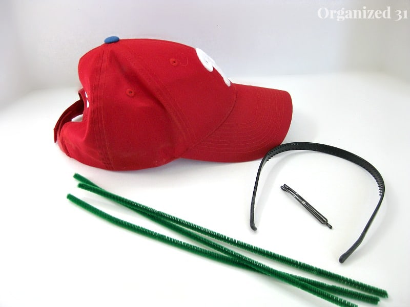 red baseball cap, head band, hair clip and 3 green pipe cleaners on white table