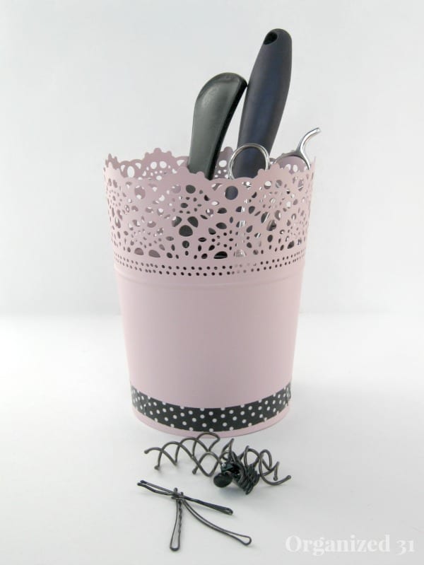 a pot wrapped in pink decorative paper  with black and white washi tape on the bottom, with scissors, a hair brush and combs in it, next to hair clips on a white table
