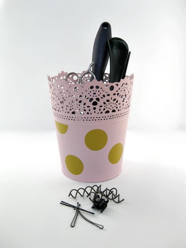 a pot wrapped in pink decorative paper with gold polka dots on it, with scissors, a hair brush and combs in it, next to hair clips on a white table
