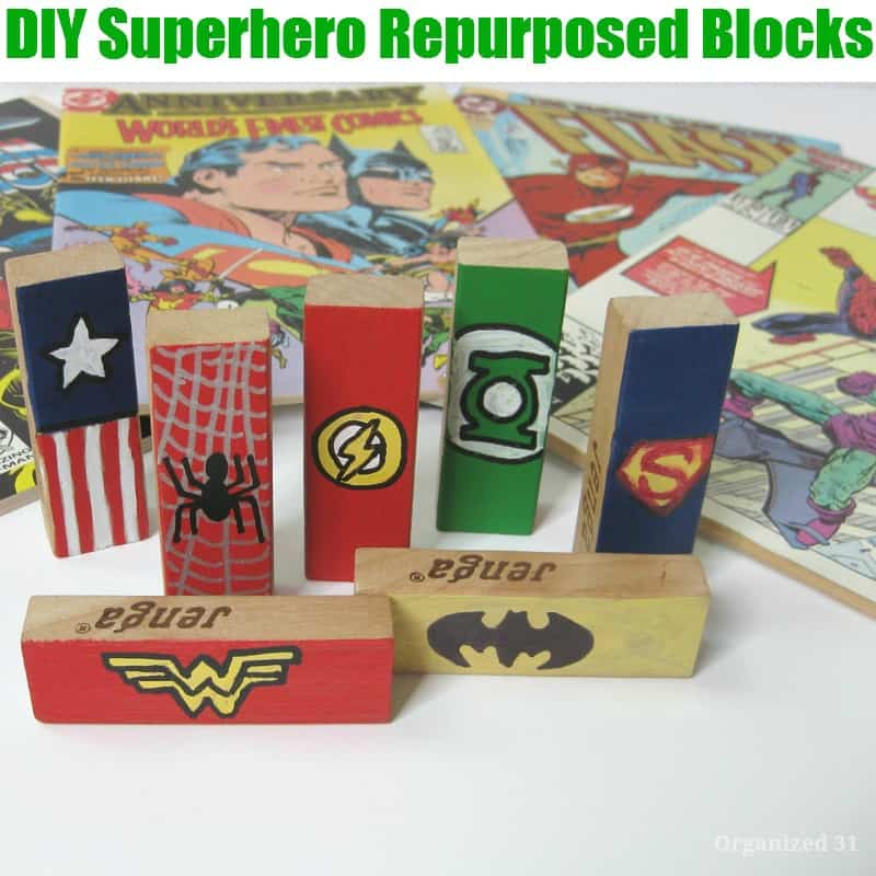 wood blocks painted with superhero logos standing up in front of a pile of comic books with title text reading DIY Superhero Repurposed Blocks