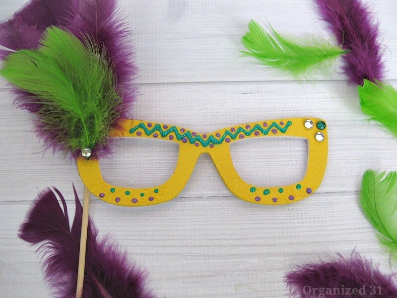 close up of yellow glasses mask decorated with green and purple puffy paint and sequins and purple and green feathers on wood table