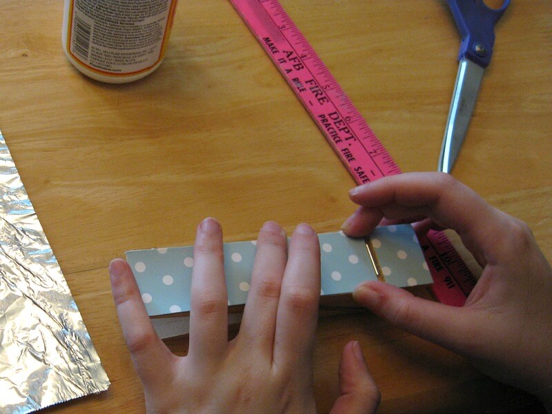 hands holding decorative paper on top of giant clothes pin with ruler, scissors and jar of decoupage glue on table