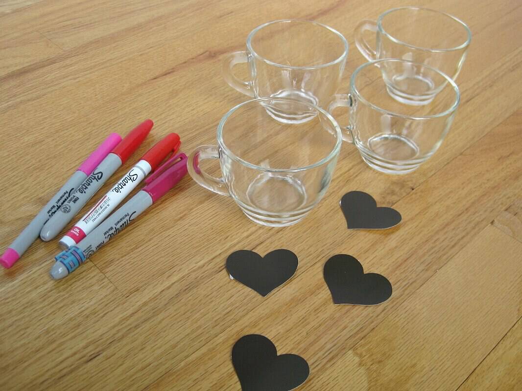 four clear cups, pile of pink and red markers and black heart stickers on table