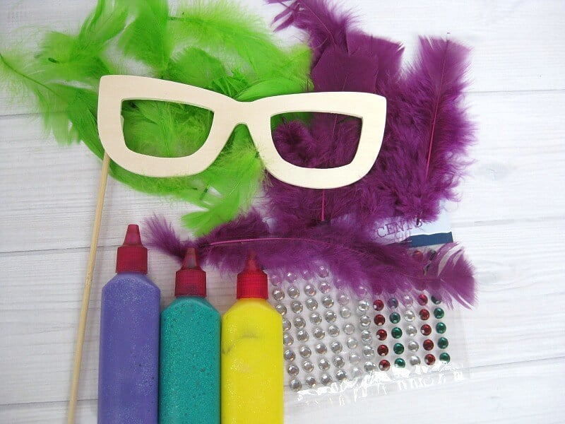 pile of green feathers, pile of purple feathers, 3 bottles of craft paint, sheets of sequins and wood glasses mask on stick