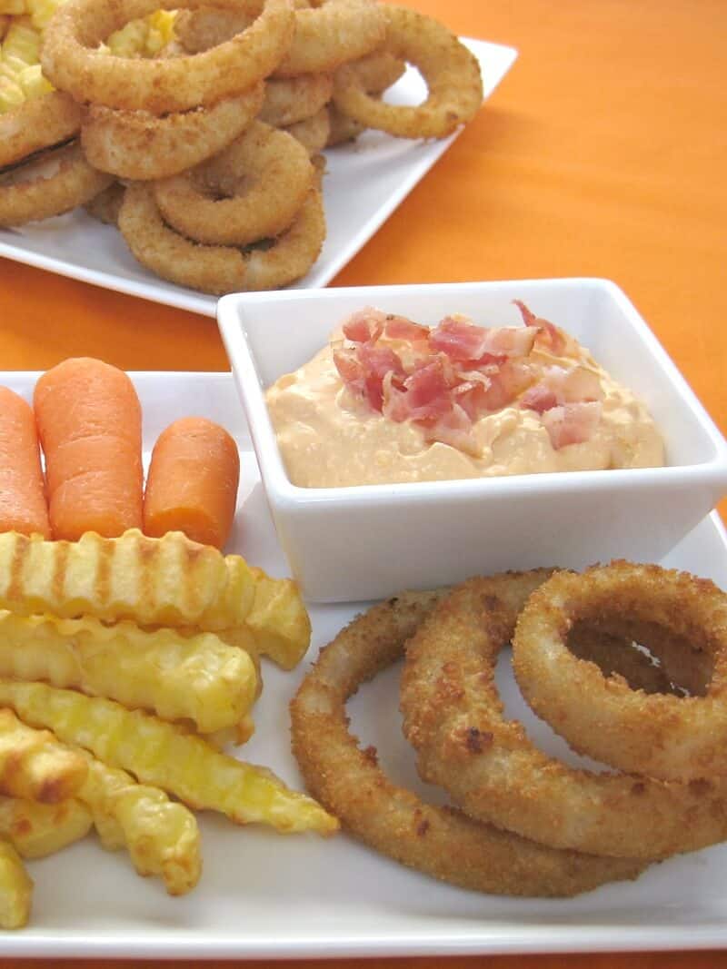 close up of white plate with celery, carrots, french fries, onion rings and a small bowl of dip with crumbled bacon with plate of french fries and onion rings in background on orange table cloth