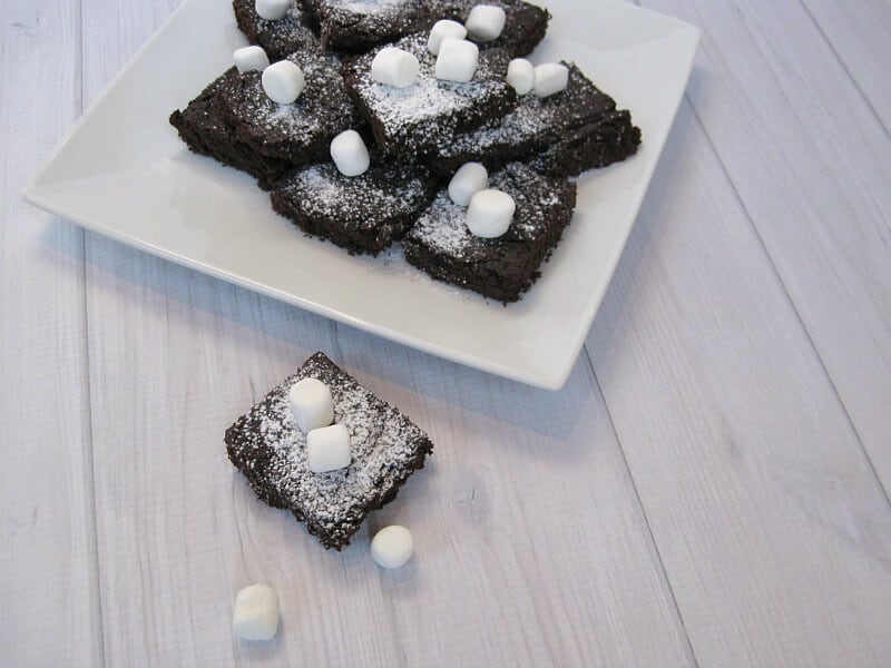 overhead view of plate of brownies with marshmallows and one brownie next to plate on white wood table