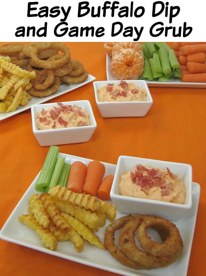close up of white plate with celery, carrots, french fries, onion rings and a small bowl of dip with crumbled bacon on orange table cloth with more food in the background