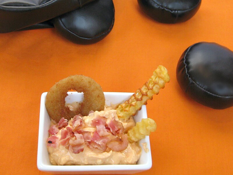 overhead view of small white bowl of dip with crumbled bacon and french fries and an onion ring standing up in the bowl with stuffed black hockey pucks in the background on orange table cloth