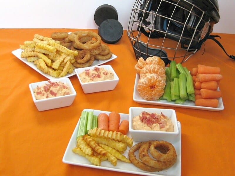 white plate with celery, carrots, french fries, onion rings and a small bowl of dip with crumbled bacon on orange table cloth with more food and a black hockey helmet in the background
