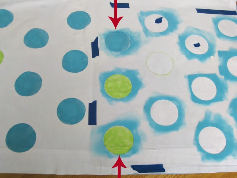 overhead view of white curtain laid out flat with green and blue stenciled polka dots and some unpainted polka dots with pieces of blue tape and red arrow overlay pointing to one blue polka dot