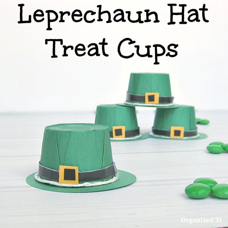 crafted green paper hats with black strap and buckle 3 stacked in pyramid and one in front and green candies scattered on white wood table