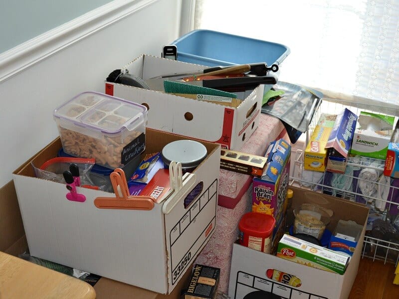 several boxes with boxes of crackers adn food, kitchen wrap and bags and cereals stacked in corner of room by window