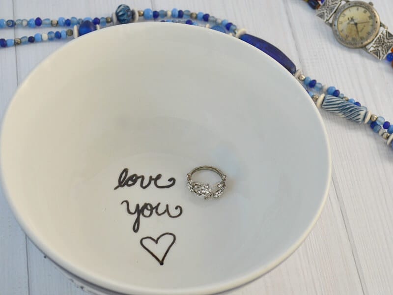 interior of bowl with black "love you" and hear and one silver ring on white wood table with blue necklace and watch in background