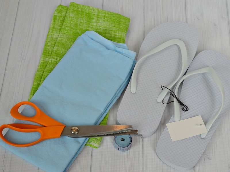 folded blue and green fabric, white rubber flip flops, orange handled scissors and blue measuring tape on white wood table
