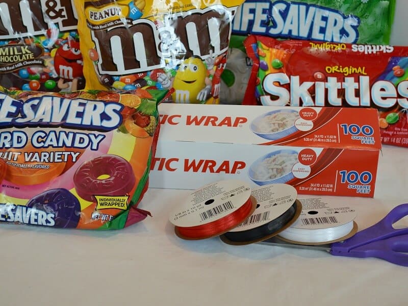 brightly colored bags of candy with box of plastic wrap, rolls of red, black and white ribbon with purple handled scissors