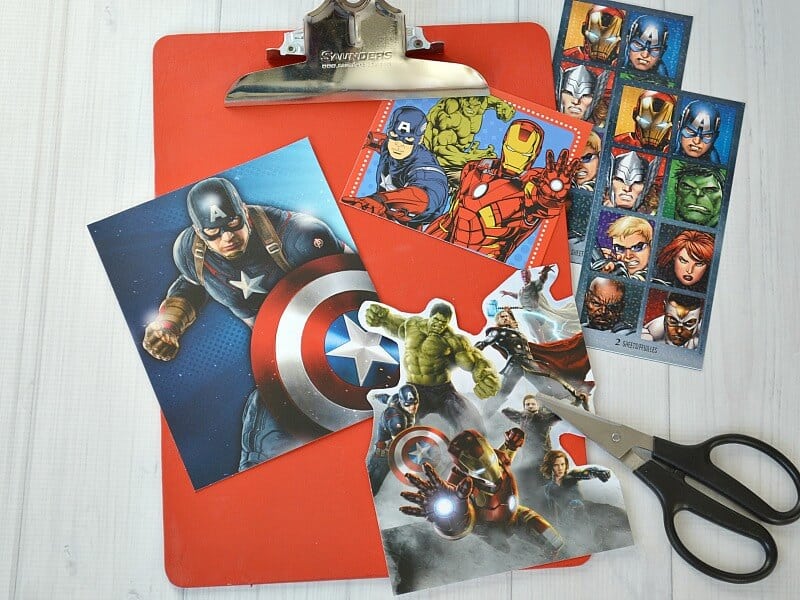 overhead view of red clipboard with sheets of superhero stickers on table and superhero cards on top of clipboard with pair of black handled scissors
