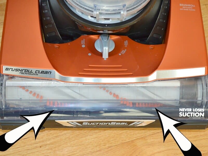 front of orange vacuum with white and black arrow overlays pointing to vacuum brush with no hair tangled