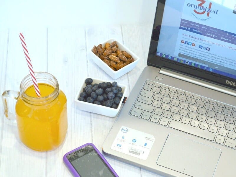 overhead view of mason jar glass with red and white straw and orange drink next two 2 small bowls of blueberries and nuts next to laptop and cellphone