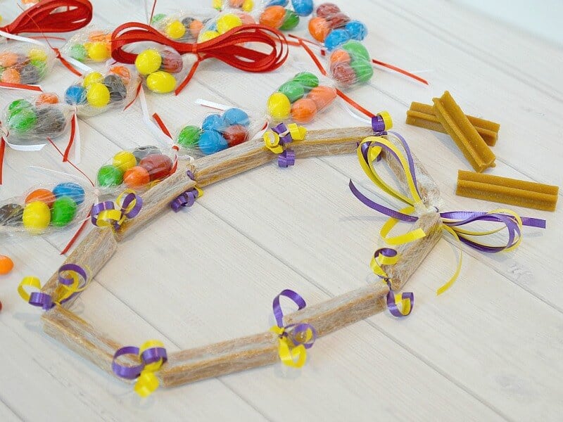 dog treat lei with purple and yellow ribbons laying on white wood table next to colorful candy lei