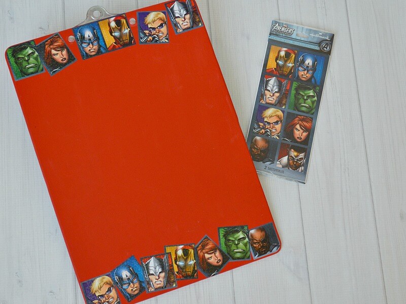 red clipboard with square superhero stickers in a line at the top and bottom of clipboard with package of stickers on table next to it