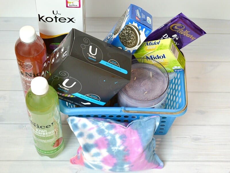 overhead view of blue basket filled with cookies, candy, tampons and a purple and blue rice heat pack