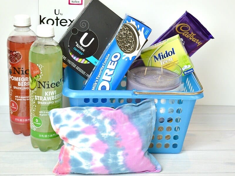 blue basket filled with cookies, candy, tampons, a candle and a purple and blue rice heat pack with 2 bottles of juice on white wood table