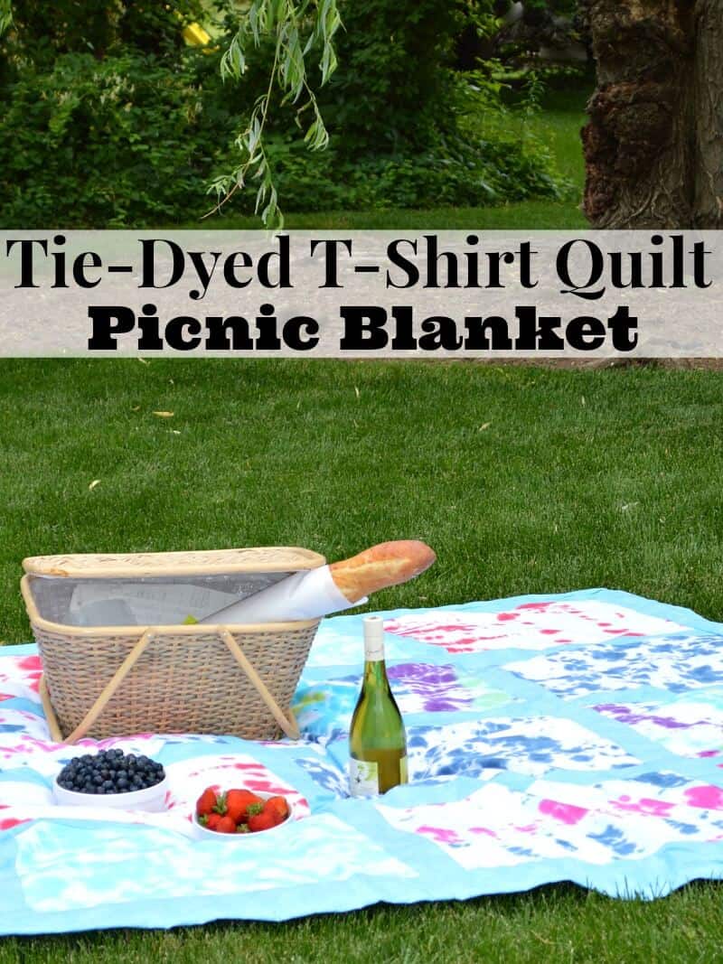 picnic basket with loaf of bread, bottle of wine and 2 bowls of fruit of colorful tie-dyed blanket on grass