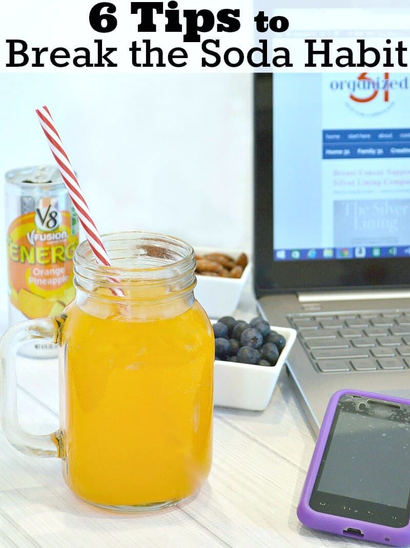 mason jar glass with orange drink and red and white straw next to laptop and cell phone with 2 small white bowls of blueberries and nuts and silver can of energy fruit drink