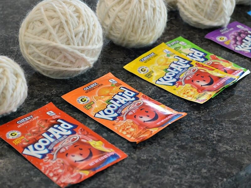 row of 4 balls of white yarn with packets of Kool-Aid on black counter 