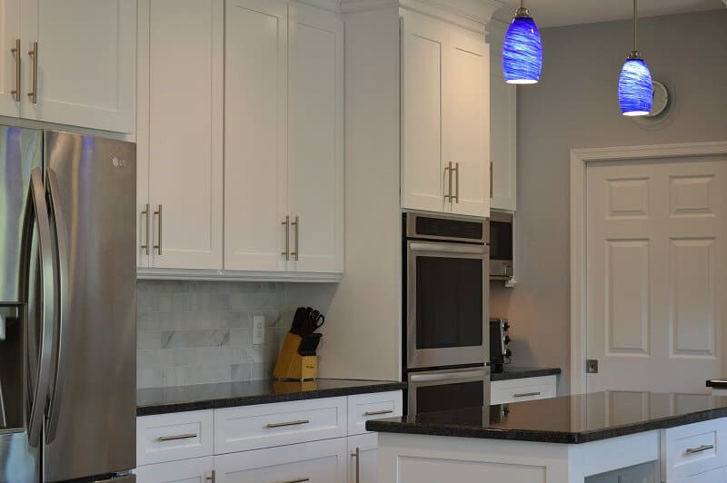 Double oven and closed pocket door in a modern white kitchen with black granite counters, white marble backsplash and blue hanging lights