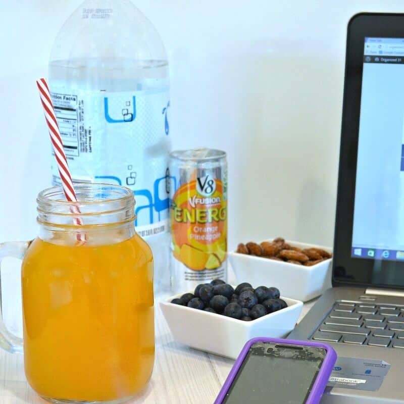 jar with orange drink and red and white straw next to cell phone and laptop with small bowls of blueberries and nuts and a drink can 