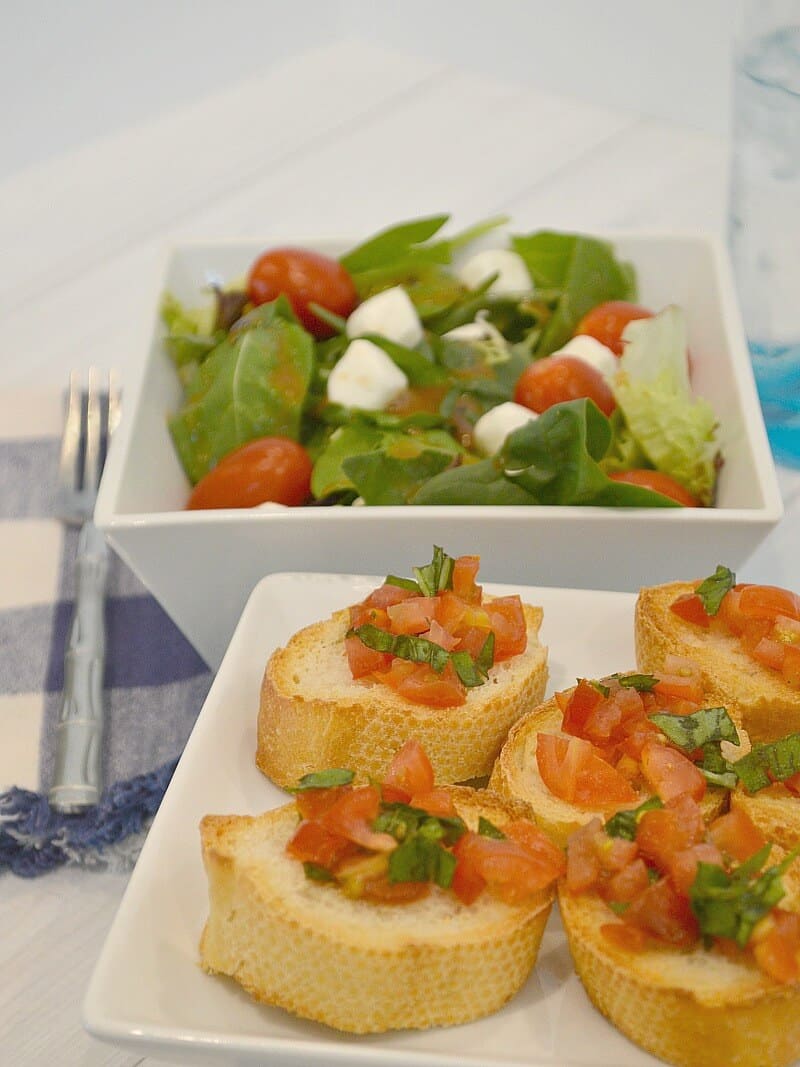 white plate of bruschetta on bread slices with white bowl of salad, blue and white napkin and fork in background