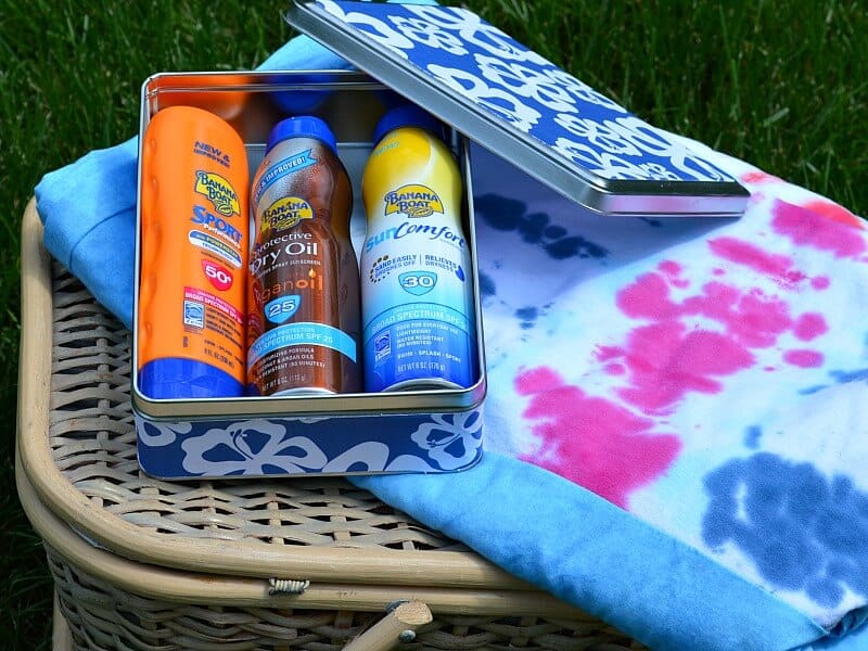 3 bottles of sunscreen in metal box with decorative paper sitting on colorful quilt on a picnic basket 