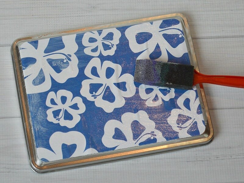 red handled sponge brush adding decoupage glue to blue and white floral paper on metal box lid