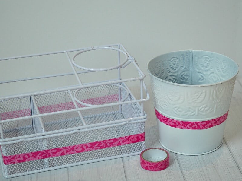 white wire basket with pink washi tape stripe next to white bucket with pink washi tape stripe and roll of washi tape on white wood table