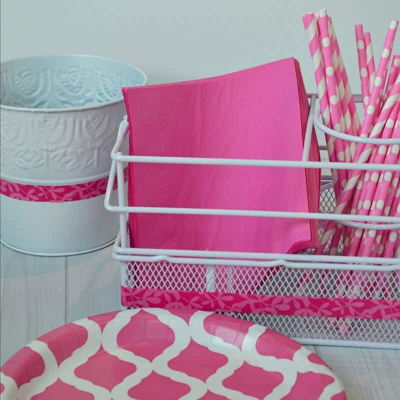 white bucket with pink stripe, white basket holding pink napkins and pink polka dot straws next to stack of pink plates on white wood table