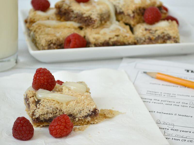 one light brown baked bar with raspberries and glaze on napkin with school worksheet and pencil, glass of milk and plate of baked browns in background