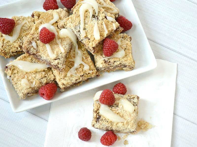overhead view of light brown baked bars with icing and fresh raspberries on white plate and one on a napkin by plate