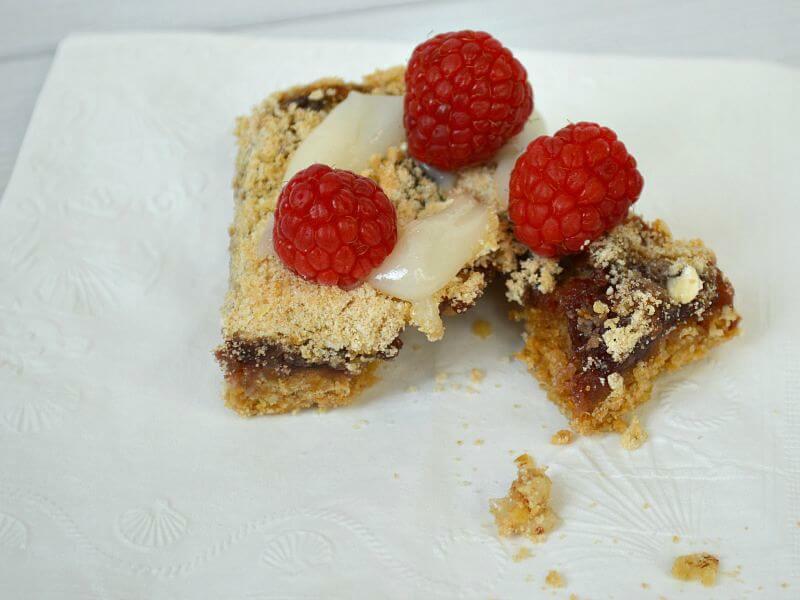 light brown baked bar with jam filling with glaze on top and fresh raspberries broken open with crumbs on white napkin